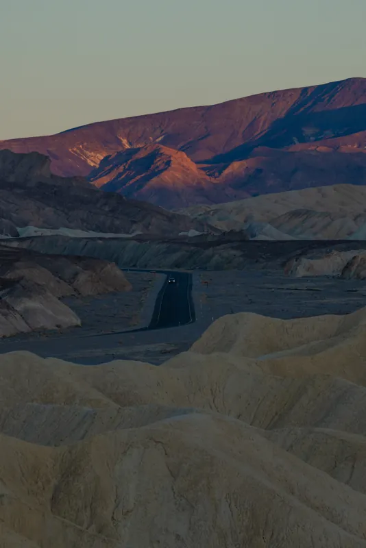 A road winding through pale badlands toward a mountain lit violet at dusk