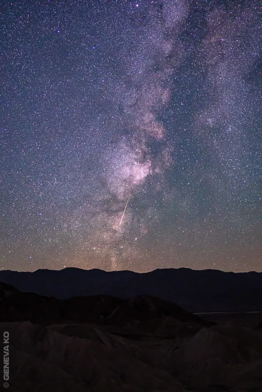The Milky Way arching over dark desert hills, a meteor streaking through
